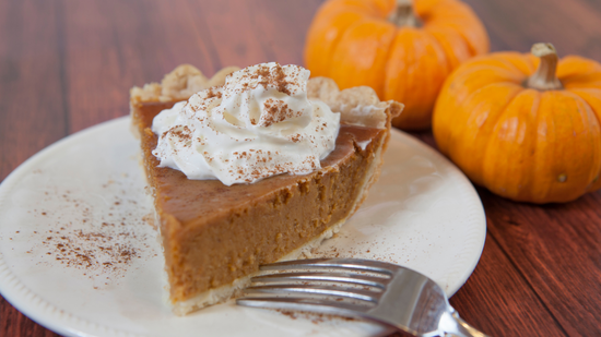 Slice of pumpkin pie with whip cream. Two small pumpkins are on the table in the back.